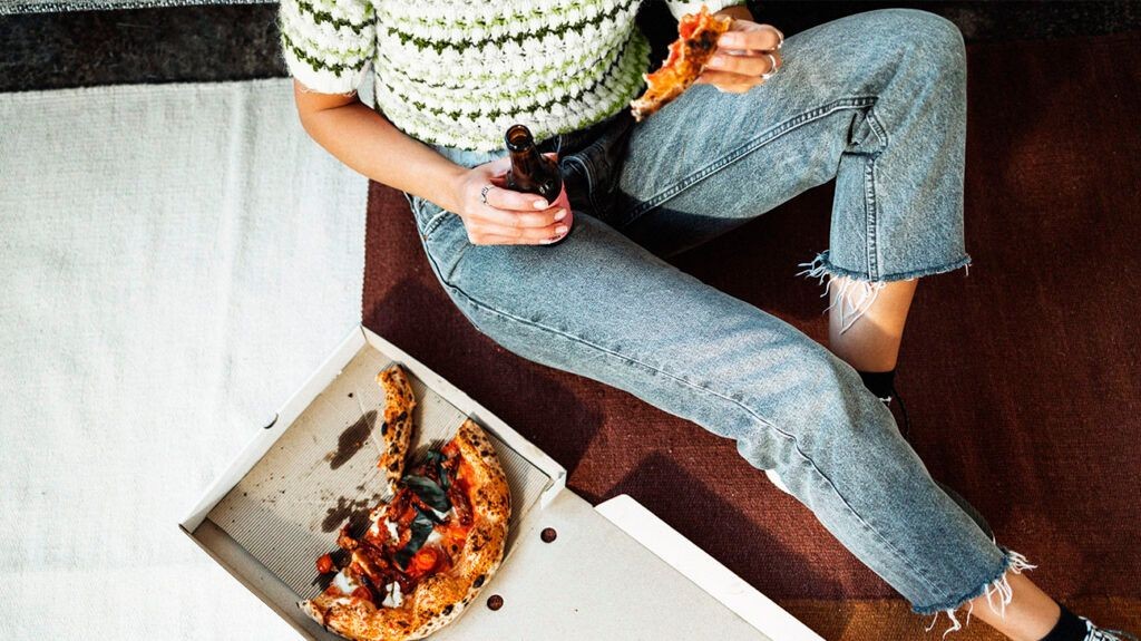 A person experiencing flatulence while eating pizza and drinking from a bottle, illustrating common triggers for intestinal gas.