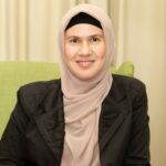 A smiling woman wearing a beige hijab and a black undercap.