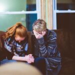 A woman prays with her head bowed on a couch, symbolizing grief and the comfort found in faith, relevant to the Bible verse 'Jesus wept'.