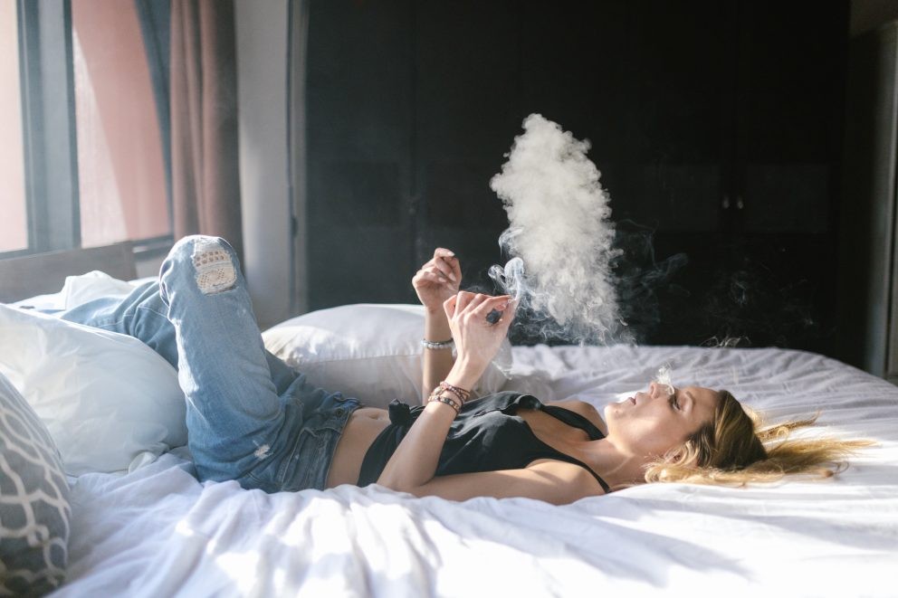 A woman wearing tank top and denim jeans lying on the bed while smoking