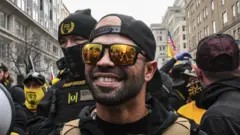 Getty Images Former Proud Boys leader Enrique Tarrio is shown smiling while wearing mirrored sunglasses and back and yellow protective clothing. He is standing outside Harry