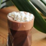 Hands holding a mug of hot chocolate with marshmallows, steam rising, against a blurred winter background.