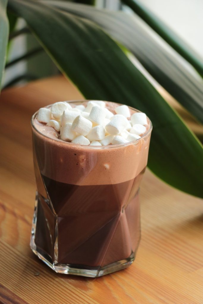 Hands holding a mug of hot chocolate with marshmallows, steam rising, against a blurred winter background.