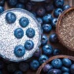 Overhead view of chia seed pudding with almond milk and blueberries, next to coconut shells holding dry chia seeds and blueberries