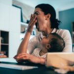 Tired mother with a baby trying to work on laptop at home and yawning