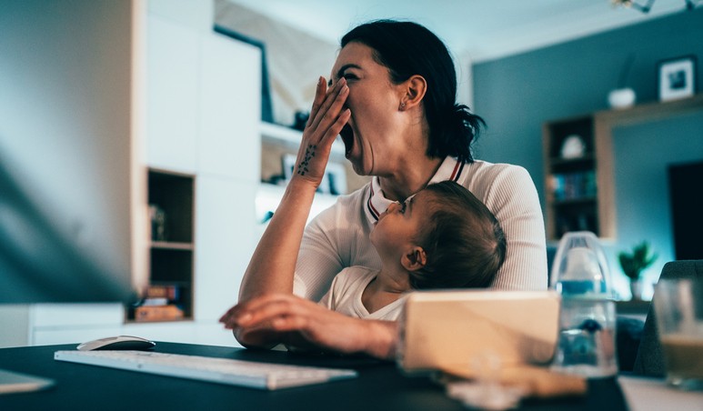 Tired mother with a baby trying to work on laptop at home and yawning