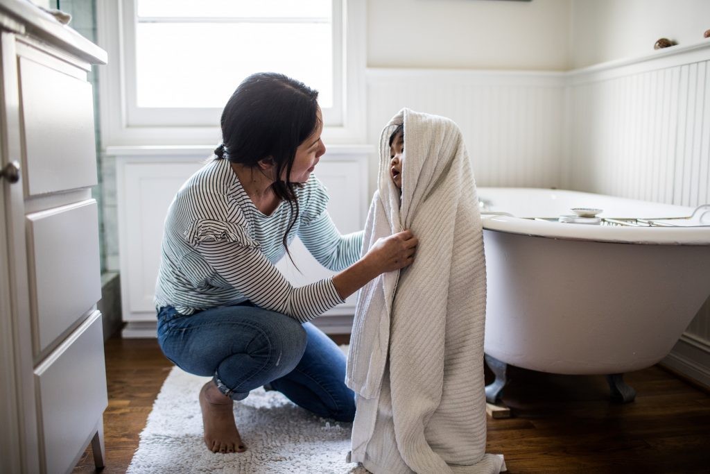 Woman assisting a young child wrapped in a white towel stepping out of a bathtub